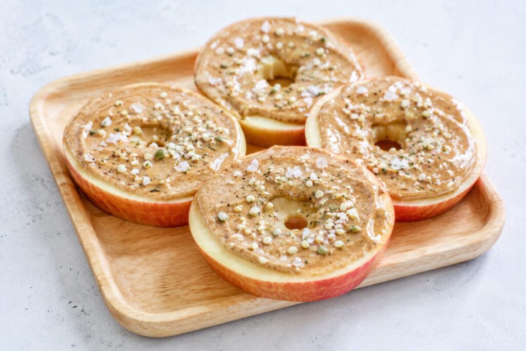 Apple rings topped with almond butter, hemp seeds, and flaky sea salt on a wooden tray.