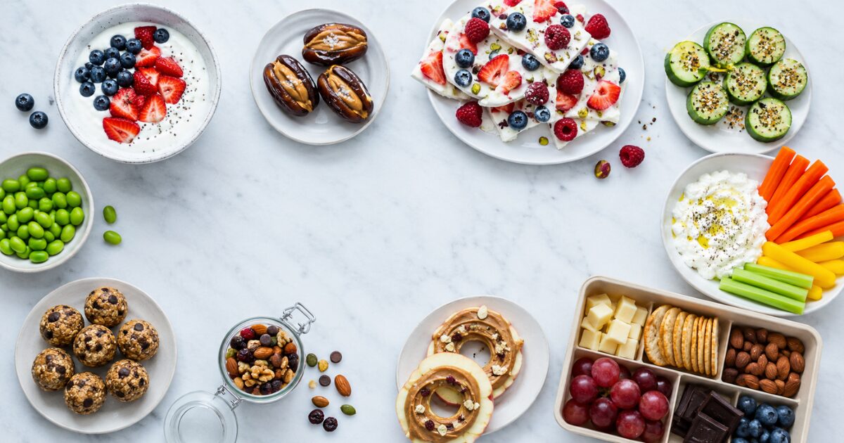Bright overhead spread of simple healthy snacks including yogurt with berries, stuffed dates, cucumber bites, energy bites, trail mix, and a divided snack box.