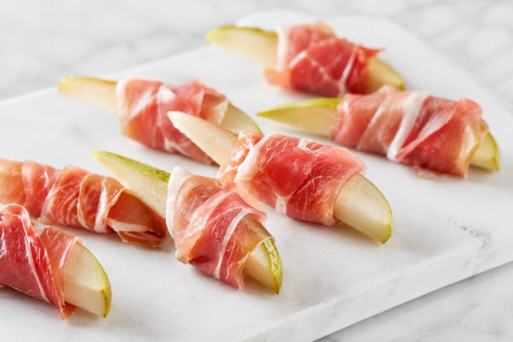 Pear wedges wrapped with thin prosciutto ribbons on a white marble board.