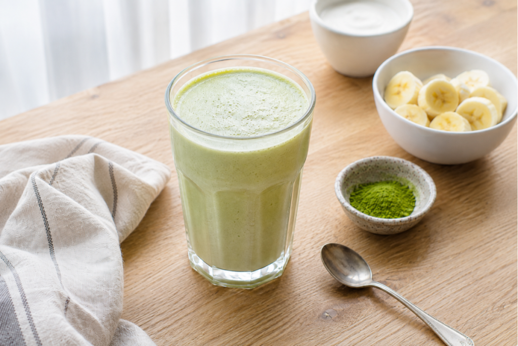 Creamy soft-green matcha vanilla protein smoothie beside banana slices and matcha powder.